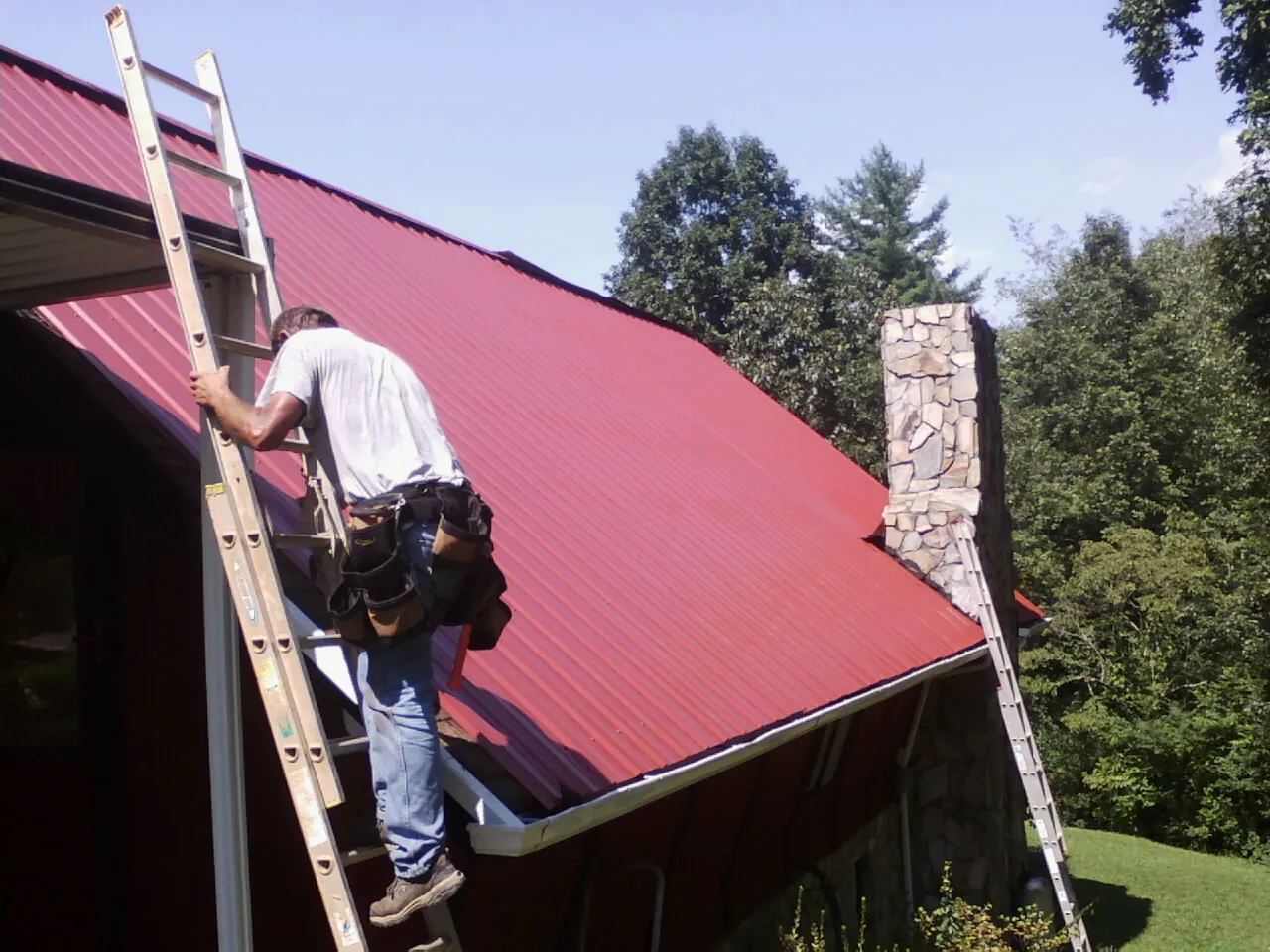 Roofer on a ladder beside a red metal roof and stone chimney — Soffit & Fascia Repair by Roofing Brothers in Jeffersonton